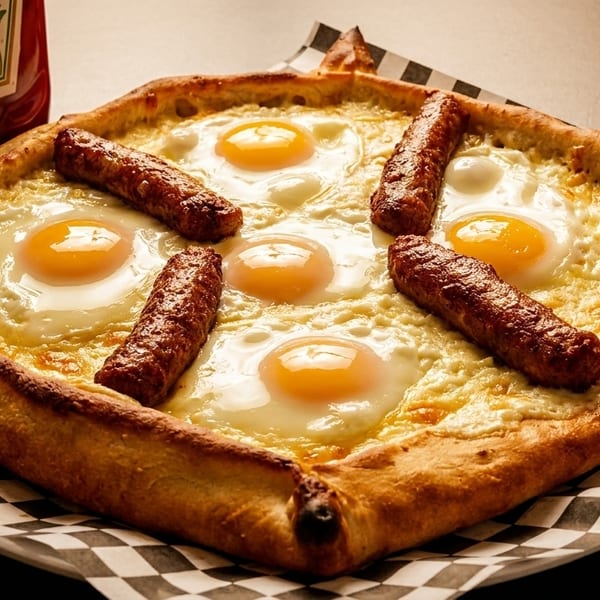 Breakfast pizza with five fried eggs and five sausage links, served on checkered paper with ketchup.