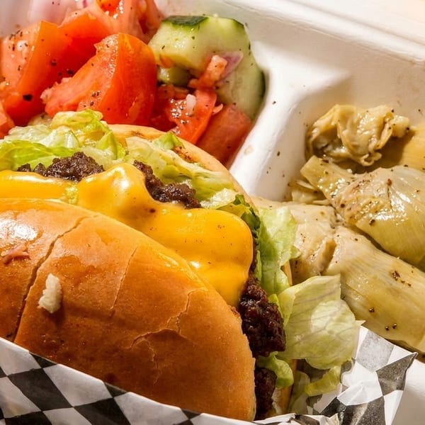 Cheeseburger with lettuce, tomato salad, and artichoke hearts in a white takeout container.